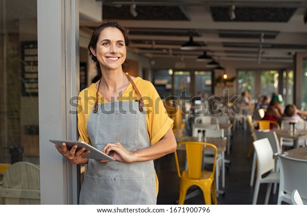 Portrait of a happy waitress standing at\
restaurant entrance holding digital tablet. Happy mature woman\
owner in grey apron standing at coffee shop entrance leaning while\
looking away with copy\
space.