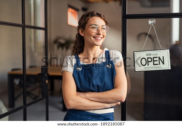 Portrait of happy waitress standing at restaurant\
entrance. Portrait of young business woman attend new customers in\
her coffee shop. Smiling small business owner showing open sign in\
her shop.