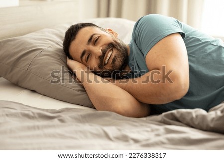 Portrait of happy well-slept man lying in bed, looking and smiling at camera, resting in bedroom in the morning, free copy space