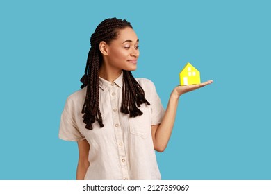 Portrait Of Happy Woman With Black Dreadlocks Standing With Paper House On His Palm And Smiling, Home Purchase, Wearing White Shirt. Indoor Studio Shot Isolated On Blue Background.