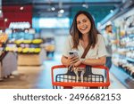 Portrait of happy woman buying groceries in supermarket and looking at camera. Purchasing Goods with Smartphone at Grocery Store. Female customer shopping with smartphone checklist