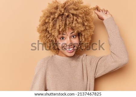 Portrait of happy woman curls hair has natural curls shiny well cared hair after washing with shampoo smiles toothily dressed in casual jumper isolated over beige background. Human emotions. Stock photo © 