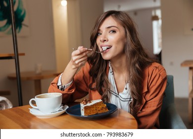 Portrait of a happy woman eating a piece of cake and drinking coffee while sitting at the table in a cafe indoors - Powered by Shutterstock