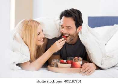 Portrait Of A Happy Woman Feeding Strawberry To Man For Breakfast. Young Smiling Couple Eating Breakfast On Bed. Happy Young Couple Enjoying Their Breakfast At Home. 