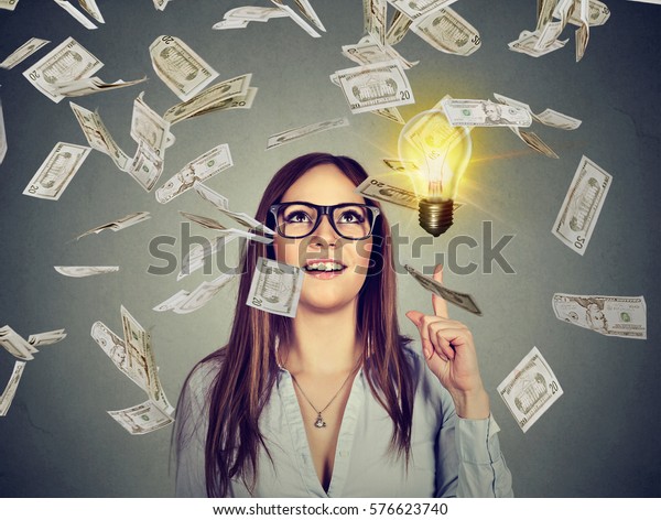 Portrait happy woman in glasses has a successful idea under money rain isolated on gray wall background with bright light bulb above head 