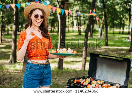 portrait of happy woman in hat and sunglasses with grilled vegetables on plate showing ok sign in park