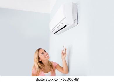 Portrait Of A Happy Woman Holding Remote Control In Front Of Air Conditioner At Home