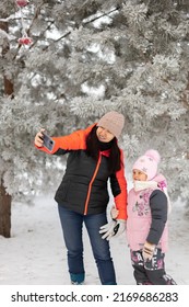 Portrait Of Happy Woman Of Middle Age Making Selfie Of Herself And Daughter Smiling Standing On Snowy Ground Near Trees Both Looking At Phone. Having Fun Outside In Winter. Family Time.