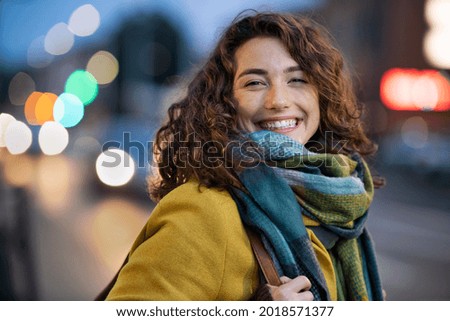Portrait of happy woman on city street looking at camera. Beautiful girl standing outdoors in winter evening with a toothy smile. Cheerful university student wearing coat with woolen scarf at dusk.