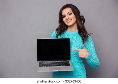 Portrait Of A Happy Woman Pointing Finger On Blank Laptop Computer Screen Over Gray Background And Looking At Camera