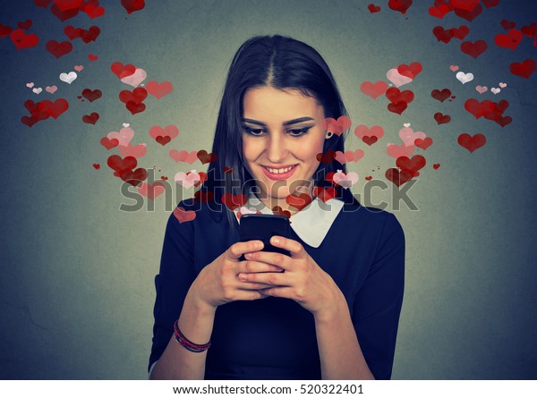 Portrait happy woman sending love sms text message on mobile phone with red hearts flying away from screen isolated on gray wall background. Human emotions feelings 