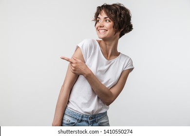 Portrait of happy woman with short hair in basic t-shirt rejoicing and pointing finger at copyspace isolated over white background