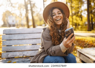 Portrait of happy woman with smartphone on bench in sunny autumn park. Woman enjoys nature while talking on the phone. Concept of relaxation, recreation