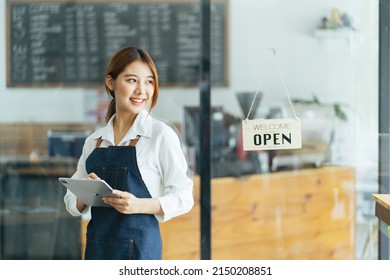 Portrait Of Happy Woman Standing At Doorway Of Her Store. Cheerful Mature Waitress Waiting For Clients At Coffee Shop. Successful Small Business Owner In Casual Wearing Blue Apron Standing At Entrance