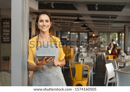 Portrait of happy woman standing at doorway of her store. Cheerful mature waitress waiting for clients at coffee shop. Successful small business owner in casual wearing grey apron standing at entrance