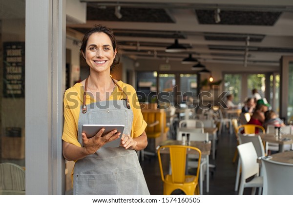 店の戸口に立つ幸せな女性のポートレート 元気な成熟したウエートレスが喫茶店で客を待っている 入り口に立つグレーのエプロンをカジュアルに着用し スモールビジネスの成功者 の写真素材 今すぐ編集