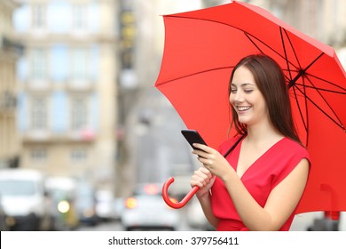 Portrait of a happy woman wearing red blouse texting on a smart phone under an umbrella in a rainy day in the street of an old town - Powered by Shutterstock