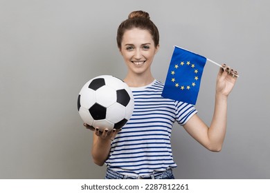 Portrait of happy woman wearing striped T-shirt holding in hands flag of europe union and football ball, supporting favourite team on championship. Indoor studio shot isolated on gray background. - Powered by Shutterstock