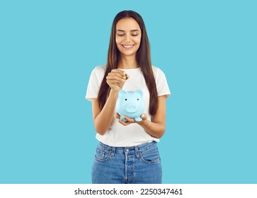 Portrait of happy woman who puts coin in piggy bank, saving money for purchase she dreams of. Young woman in casual clothes holding piggy bank in shape of pig isolated on light blue background.