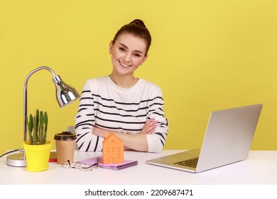 Portrait Of Happy Woman At Workplace With Little Paper House Looking At Camera With Smile, Dreaming About Her Own Apartment. Indoor Studio Studio Shot Isolated On Yellow Background.