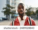 Portrait of happy young adult African man hipster standing on city street outdoors looking at camera. Smiling cool generation z Black hipster guy wearing red shirt headphones posing for close up photo