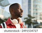 Portrait of happy young adult African man hipster standing on city street outdoors looking away dreaming. Smiling cool gen z Black hipster guy traveler wearing red shirt headphones backpack. Close up