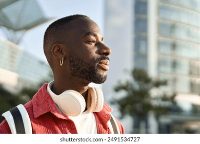 Portrait of happy young adult African man hipster standing on city street outdoors looking away dreaming. Smiling cool gen z Black hipster guy traveler wearing red shirt headphones backpack. Close up - Powered by Shutterstock