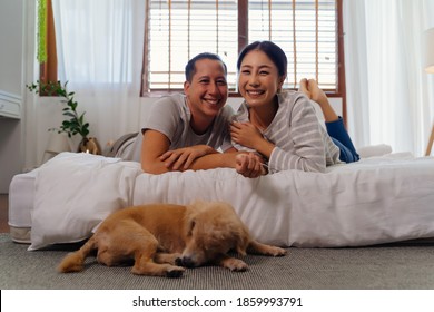 Portrait Of Happy Young Adult Asian Couple Looking At Camera On Bed Together With Dog Pet In Bedroom Interior Scene. 30s Mature Husband And Wife Smiling With Happiness. Happy Relationship Life Concept