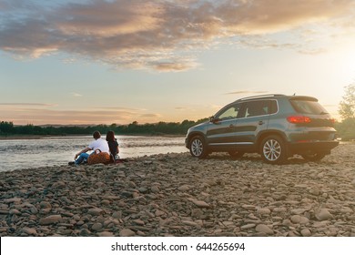 Portrait of happy young adult couple with dog on roadtrip. Man sitting on plaid with woman and watching sunset near river. Outdoor picnic concept.