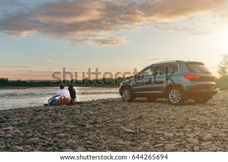 Portrait of happy young adult couple with dog on roadtrip. Man sitting on plaid with woman and watching sunset near river. Outdoor picnic concept.