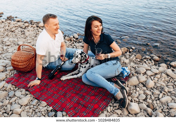 Portrait of\
happy young adult couple with dog on roadtrip. Man sitting on plaid\
with woman. Outdoor picnic\
concept.