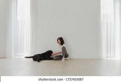 Portrait of a happy young adult in good mood sitting on a table at home with crossed legs. Happy housewife caresses a black labrador with pleasure. Copy space, family concept
