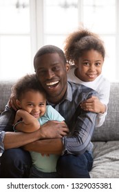 Portrait Of Happy Young African American Family Of Three Relax At Home Sit On Couch Hugging Posing For Picture, Smiling Black Dad Play With Small Kids, Have Fun Laughing And Embracing On Sofa