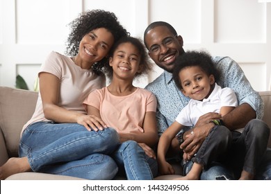 Portrait Of Happy Young African American Family With Little Kids Sit Relax On Couch Cuddling, Smiling Black Parents Rest On Sofa Hug Preschooler Children Posing For Picture At Home Together