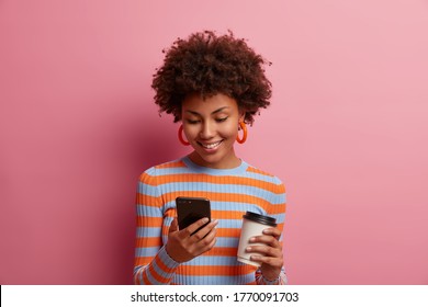 Portrait of happy young African American woman types message, uses new mobile application, searches web and drinks takeaway coffee, has positive expression, wears striped jumper, stands indoor