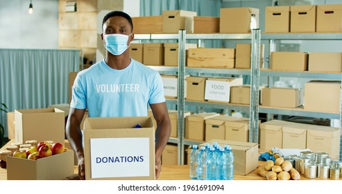 Portrait Of Happy Young African American Handsome Guy Volunteer Standing In Warehouse Storage Holding Donations Box In Hands, Looking At Camera In Good Mood During Covid Pandemic, Charity Concept