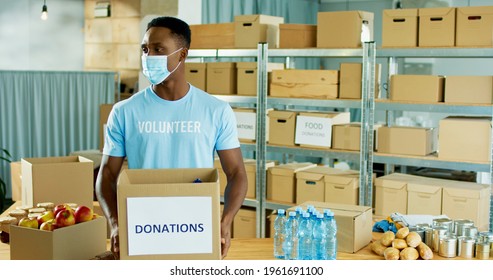 Portrait Of Happy Young African American Handsome Guy Volunteer Standing In Warehouse Storage Holding Donations Box In Hands, Looking At Camera In Good Mood During Covid Pandemic, Charity Concept