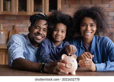 Portrait Of Happy Young African American Family With Teen Daughter Put Coin Save Money In Piggybank. Smiling Economical Ethnic Parents With Child Feel Provident Make Financial Investment For Future.