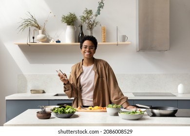 Portrait Of Happy Young African American Woman In Eyeglasses Holding Cellphone In Hands, Looking At Camera Distracted From Cooking Healthy Food In Modern Kitchen, Web Surfing Tasty Recipe Online.