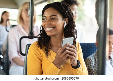 Portrait of happy young African American woman passenger sitting on bus seat with wireless headphones using smartphone while moving in the modern tram, enjoying trip at public transport, looking away - Powered by Shutterstock