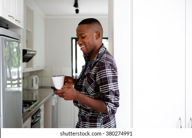 Portrait Of Happy Young African Guy Standing In Kitchen Holding A Cup Of Coffee And Looking At Mobile Phone