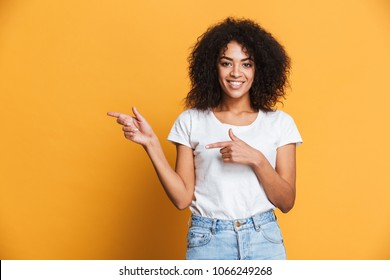 Portrait of a happy young african woman pointing fingers away at copy space isolated over yellow background
