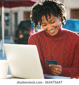 Portrait Of Happy Young African Woman Holding A Credit Card And Shopping On Internet. Girl Using Laptop For Online Banking At Cafe. Smiling Woman Using Laptop And Credit Card For Online Payment.
