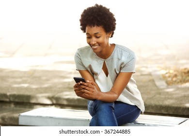 Portrait Of Happy Young African Woman Sitting Outside On Bench Reading A Text Message On Her Mobile Phone