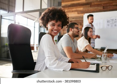 Portrait Of Happy Young African Woman Sitting At A Business Presentation With Colleagues In Boardroom. Female Designer With Coworkers In Conference Room.