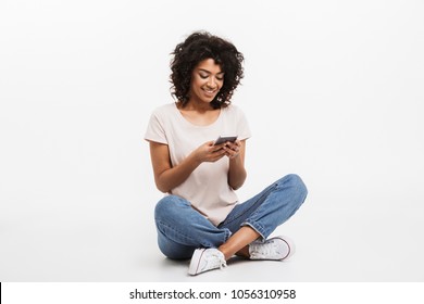 Portrait of happy young afro american woman using mobile phone while sitting on a floor with legs crossed isolated over white background