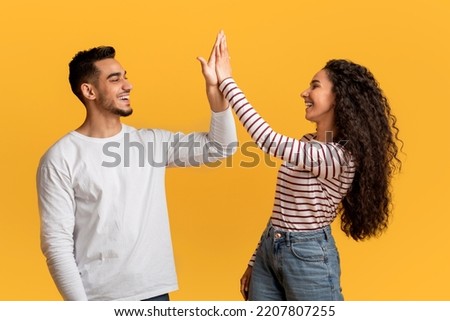 Portrait Of Happy Young Arab Couple Giving High Five To Each Other, Cheerful Middle Eastern Man And Woman Enjoying Teamwork, Celebrating Common Success While Standing Over Yellow Studio Background