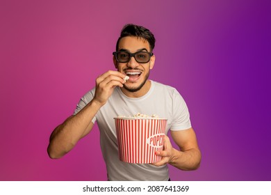 Portrait Of Happy Young Arab Guy In 3d Glasses Eating Popcorn From Bucket, Watching Movie In Cinema, Neon Light. Cheerful Middle Eastern Man Enjoying Good Film, Having Yummy Snack