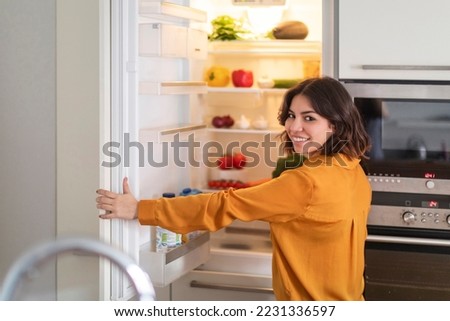 Portrait Of Happy Young Arab Woman Opening Fridge In Kitchen, Beautiful Middle Eastern Female Standing Near Refridgerator Full Of Fruits And Vegetables And Smiling At Camera, Enjoying Healthy Food