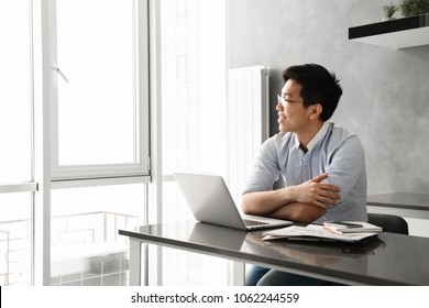 Portrait Of A Happy Young Asian Man Working On Laptop Computer While Sitting At The Table With Paperwork At Home